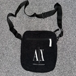 Svart axelväska från Armani Exchange - Snygg svart axelväska från Armani Exchange med justerbar rem och dragkedja. Perfekt för att bära dina nödvändigheter med stil. Väskan har en enkel och modern design med logotyp framtill.