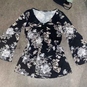 Svart blommig blus - Snygg svart blus med blommigt mönster i vitt och beige. Den har en v-ringad hals och långa ärmar med en lätt utsvängd form. Perfekt för en stilfull look.