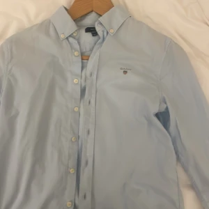 Ljusblå skjorta från GANT - Säljer en stilren ljusblå skjorta från GANT med broderad logga på bröstet. Skjortan har knappar framtill och en klassisk krage. Perfekt för en snygg och avslappnad look.