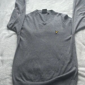Grå tröja med v-ringning från Lyle & Scott - Säljer en stilren grå tröja från Lyle & Scott med v-ringning och deras ikoniska logga på bröstet. Tröjan har långa ärmar och ribbstickade muddar vid ärmslut och nederkant. Perfekt för en avslappnad men snygg look.