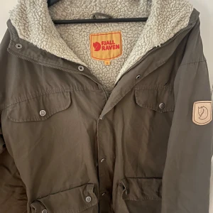 Brun jacka från Fjällräven - Säljer en mörk grönjacka från Fjällräven med mjukt fleecefoder och praktiska fickor. Jackan har knappar och dragkedja framtill samt en broderad logga på ärmen. Perfekt för kyliga dagar.