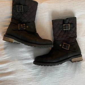 Barbour boots  - Snygga svarta boots med quiltad design och dekorativa spännen. De har en robust sula och dragkedja på sidan för enkel på- och avtagning. Perfekta för en stilren look.