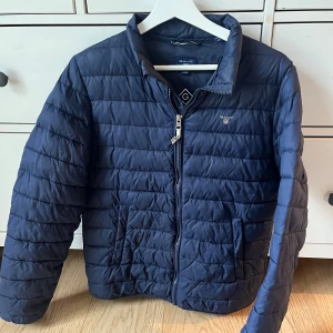 Blå quiltad jacka från Gant - Snygg blå quiltad jacka från Gant med dragkedja framtill. Jackan har en klassisk krage och långa ärmar, perfekt för kyligare dagar. Den är stilren och passar till många olika outfits.