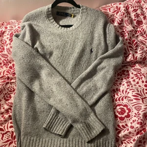Grå stickad tröja från Polo Ralph Lauren - Säljer en stilren grå stickad tröja från Polo Ralph Lauren. Tröjan har en klassisk rund halsringning och långa ärmar med ribbade muddar. Perfekt för kyligare dagar och enkel att matcha med olika outfits.