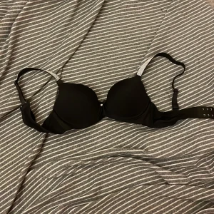 Svart bh med strassdetaljer - Elegant svart bh med glittrande strassdetaljer på axelbanden och en liten dekorativ berlock mellan kuporna. Perfekt för att lägga till lite glamour till din outfit.