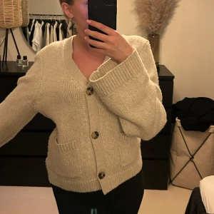 Beige stickad kofta med knappar - Mysig beige stickad kofta med stora knappar och två praktiska fickor framtill. Perfekt för kyliga dagar med sin v-ringade design och långa ärmar.