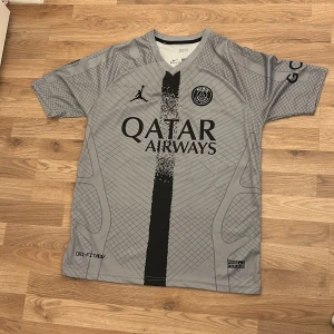 Grå PSG fotbollströja med Mbappé 7 - Snygg grå PSG fotbollströja med Mbappé 7 på ryggen. Tröjan har korta ärmar och ett stilrent mönster med klubbens emblem på bröstet. Perfekt för fans av Paris Saint-Germain och Mbappé!