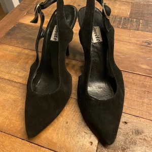 Svarta mockapumps med klack - Snygga svarta pumps i mocka med spetsig tå och elegant klack. De har en justerbar rem med guldfärgat spänne för perfekt passform. Perfekta för en utekväll eller fest! Skorna är i bra skick med lite slitage på sulan.