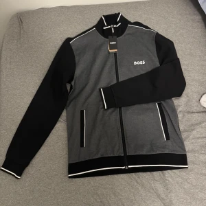 Svart och grå tröja från BOSS - Säljer en snygg svart och grå tröja från BOSS i nytt skick, helt oanvänd och som ni kan se så är lapparna fortfarande kvar. Tröjan har en dragkedja framtill och vita detaljer vid kragen och ärmsluten. Två fickor på framsidan.  Perfekt för en stilren look!