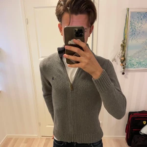 Grå stickad tröja från Polo Ralph Lauren - Säljer en snygg grå stickad hel zip  från Polo Ralph Lauren. Den har en dragkedja framtill och ribbade muddar. Perfekt för höst och vinter! Tröjan är i mycket bra skick och har en klassisk stil med en liten broderad logga på bröstet. Riktigt bra skick på denna!