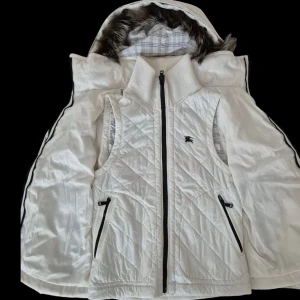 Vit jacka från Burberry Black Label - Säljer en snygg vit jacka från Burberry Black Label i storlek M. Jackan har en avtagbar huva med pälsdetaljer och ett quiltat mönster. Den har dragkedja och praktiska fickor med svarta detaljer. Perfekt för kyliga dagar!