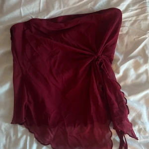 Vinröd tubtopp med knytning - Snygg vinröd tubtopp i ett lätt och luftigt material. Toppen har en elegant knytning på sidan som ger en unik touch. Perfekt för sommarens fester eller en kväll ute. Den är axelbandslös och har en lös passform som gör den bekväm att bära.