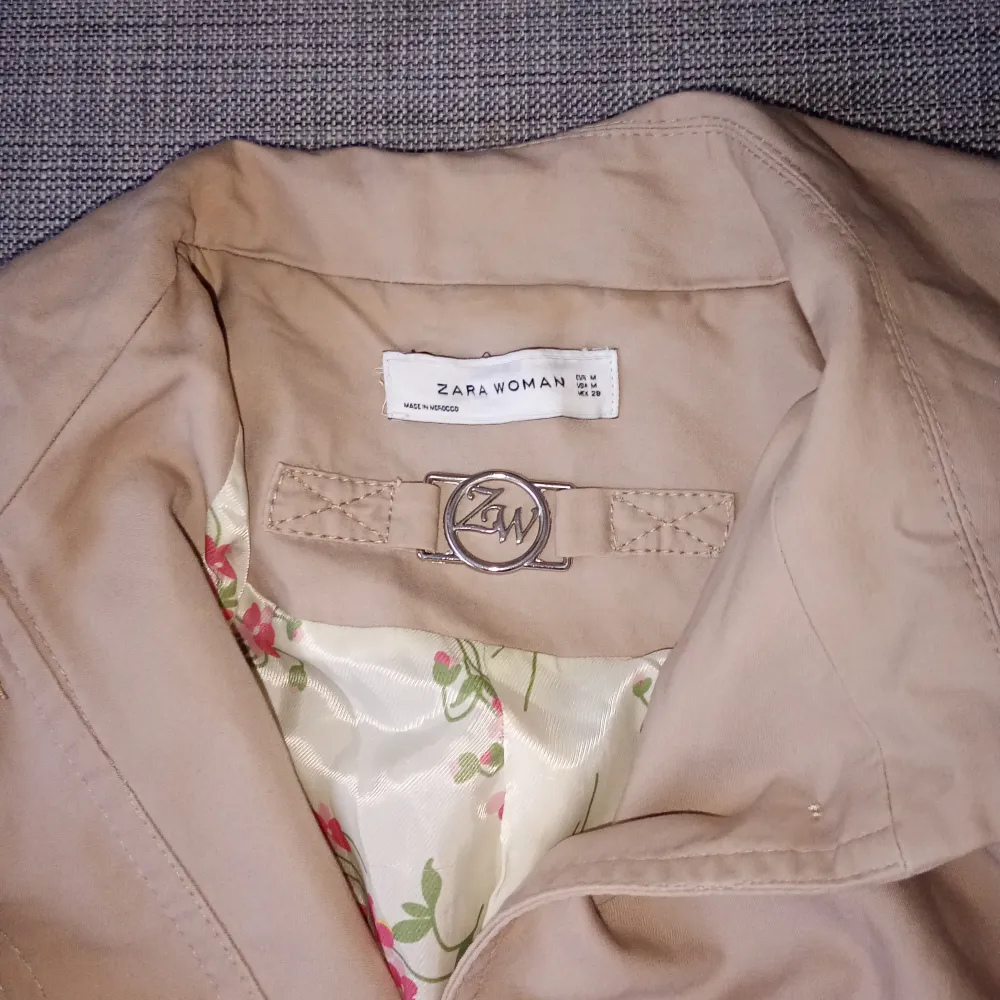 Säljer en snygg beige trenchcoat från Zara Woman i storlek M. Jackan har knappar framtill och ett fint blommigt foder som ger en extra touch. Perfekt för vår och höst!. Jackor.