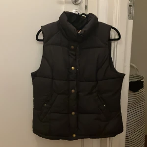 Dunväst - Säljer en svart dunväst i bra skick. Den har en hög krage och stängs med knappar framtill. Perfekt för höst och vinter när du vill hålla dig varm men ändå se stilren ut. Passar till både vardag och lite mer avslappnade tillfällen.