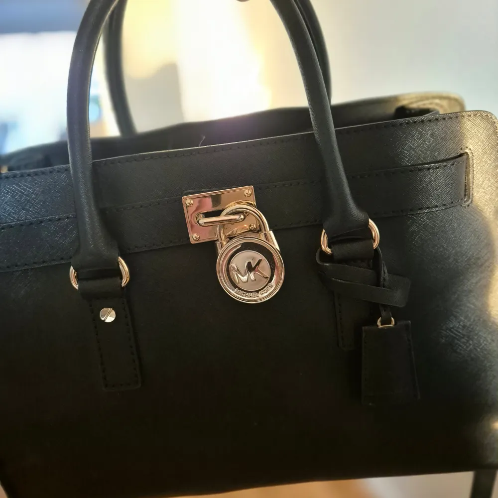 Snygg svart handväska från Michael Kors. Väskan har ett elegant lås med märkets logotyp i guld och praktiska handtag. Insidan är rymlig med flera fack och dragkedja för säker förvaring. Perfekt för både vardag och fest!. Väskor.