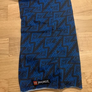 Blå och svart halsduk från LEGO Ninjago - Cool halsduk med LEGO Ninjago-tema i blått och svart. Perfekt för fans av serien! Den har ett mönster med blixtar och LEGO-klossar, vilket gör den unik och stilren. Passar perfekt för höst och vinter när du vill hålla dig varm och samtidigt visa din kärlek för Ninjago.