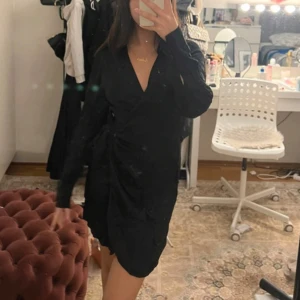 Svart omlottklänning - Snygg svart omlottklänning i mycket bra skick. Perfekt för festliga tillfällen med sin eleganta v-ringning och långa ärmar. Klänningen har en smickrande passform och är gjord i ett mjukt material som känns skönt mot huden.