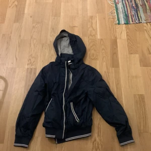 Mörkblå vindjacka från H&M - Säljer en mörkblå vindjacka från H&M i storlek 140. Jackan har en huva och dragkedja framtill. Den är fodrad med grått tyg och har ribbade muddar vid ärmslut och nederkant. Perfekt för vår och höst! Aldrig använd, helt ny 