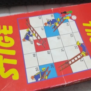 Stigespill - Barnas Favoritter - Ett klassiskt brädspel för 2-5 spelare, perfekt för barn från 4 år. Spelet har en färgglad design med stegar och brandmän som tema. Målet är att nå slutet av brädet först genom att klättra upp för stegar och undvika att glida ner. Lådan är röd med stora gula bokstäver och innehåller spelpjäser och en tärning.