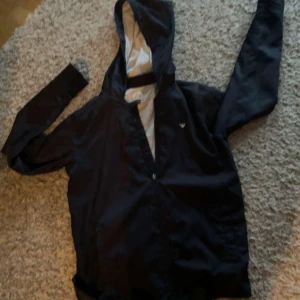 Svart jacka från Armani Junior - Säljer en snygg svart jacka från Armani Junior i storlek 14A (166 cm). Jackan har en dragkedja och en huva, perfekt för höst och vår. Den är i mycket bra skick och passar både till vardags och lite finare tillfällen.