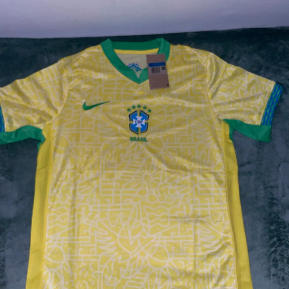 Säljer en gul Brasilien fotbollströja från Nike i nytt skick. Tröjan har ett grönt Nike-märke och Brasilien-emblem på bröstet. Den är kortärmad med gröna detaljer vid ärmsluten och har ett unikt mönster över hela tyget. Perfekt för fotbollsfans som vill visa sitt stöd för Brasilien! 💚💛. T-shirts.