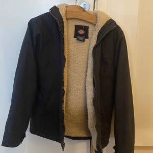 Svart jacka med teddyfoder från Dickies - Säljer en svart jacka från Dickies i mycket bra skick (har lite parfym doft fast i sig däremot). Står ingen storlek men skulle säga den är en M. Sitter Den har ett mysigt teddyfoder som håller dig varm under höst och kan nog även funka till vinter. Jackan har en dragkedja framtill och en huva för extra skydd mot kylan. Perfekt för både vardag och utomhusaktiviteter!