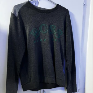 Mörkgrå sweatshirt från Hugo Boss - Säljer en snygg mörkgrå sweatshirt från Hugo Boss med grönt tryck på bröstet. Tröjan har långa ärmar och en bekväm passform, perfekt för både vardag och avslappnade tillfällen. Den är i bra skick och redo för en ny ägare!