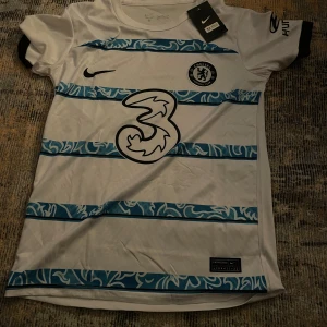 Vit och blå Chelsea fotbollströja - Säljer en vit Chelsea fotbollströja från Nike i nytt skick. Tröjan har korta ärmar och blå ränder med ett snyggt mönster. Perfekt för alla Chelsea-fans! 💙