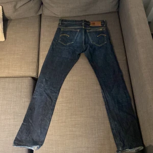 G-Star Raw jeans - Säljer ett par snygga G-Star Raw jeans i mörkblå denim. De har en klassisk femficksdesign med tydliga sömmar och en läderpatch med logga på baksidan. Jeansen har en knappgylf och ser ut att vara i bra skick. Perfekta för en casual look!