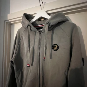 Grå hoodie från WHO's A.C.E - Säljer en snygg grå hoodie från WHO's A.C.E. Den har en dragkedja framtill och en cool logga på bröstet. På ärmen finns en detalj med texten 'ARE YOU'. Perfekt för höst och vinter, och den är i nyskick. Passar både till vardags och chill. Köpt för 1200kr på Pondus