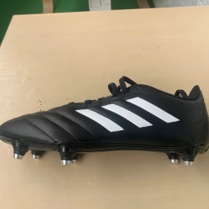 Svarta fotbollsskor från Adidas - Säljer ett par svarta fotbollsskor från Adidas med klassiska vita ränder. Skorna har snörning och är i syntetmaterial, perfekta för fotbollsträning eller match. De har en robust sula med dobbar för bra grepp på planen.
