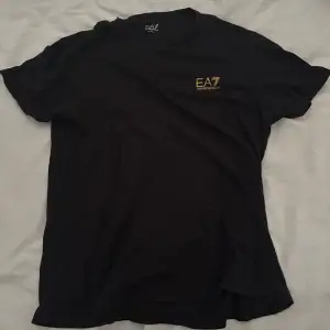 Säljer en snygg svart t-shirt från Emporio Armani EA7. Den har en stilren design med EA7-loggan i guld på bröstet och ett coolt mönster på ryggen. 