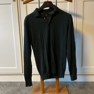 Svart långärmad pikétröja från Massimo Dutti - Säljer en stilren mörkgrön långärmad pikétröja från Massimo Dutti. Tröjan är gjord av mjuk merinoull vilket gör den perfekt för både höst och vinter. Den har en klassisk krage och knappar framtill. Perfekt för en elegant och avslappnad look.