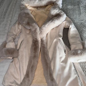 Beige pälsjacka från Garoff - Säljer en supermysig beige pälsjacka från Garoff. Den har en fluffig fuskpäls runt huvan och ärmsluten, vilket gör den perfekt för kalla vinterdagar. Jackan har en dragkedja framtill och ett bälte i midjan för en snygg passform. Insidan är också fodrad med mjuk päls för extra värme. Perfekt för både vardag och fest!  Den är i storlek XL men passar även L strålande.