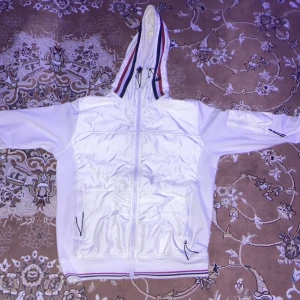 Vit jacka från Moncler - Säljer en snygg vit jacka från Moncler med dragkedja och huva. Jackan har Moncler-logotypen på ärmen och randiga detaljer i rött och blått på huvan och nederkanten. Perfekt för en stilren look.