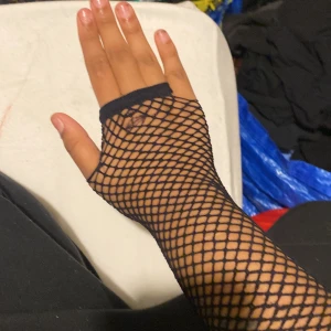 Svarta nätmönstrade handskar (Fishnet Gloves) - Snygga svarta nätmönstrade handskar som ger en edgy look. Perfekta för att komplettera en alternativ stil eller för en utekväll. De är fingerlösa och sträcker sig upp till underarmen.