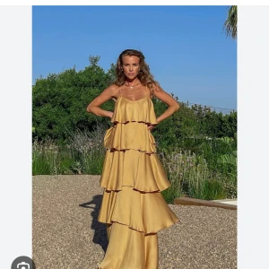 Molly rustas x NA-KD  - Säljer denna fantastiska guldiga långklänning med flera lager av volanger. Klänningen har smala axelband och en luftig passform, perfekt för sommarn. Strl 34!