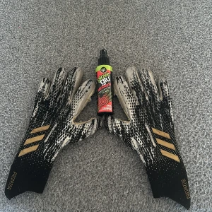  Svarta och vita Adidas predator  handskar med guldmönster - Säljer ett par coola Adidas handskar i svart och vitt med guldiga detaljer. Perfekta för fotbollsspelare som vill ha bra grepp och stil. Inkluderar en flaska Glove Glu för extra greppförstärkning.