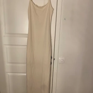 Beige långklänning med knappar - Säljer en stilren beige långklänning med smala axelband och knappar på sidan. Perfekt för en elegant och minimalistisk look. Klänningen har en enkel och tidlös design.