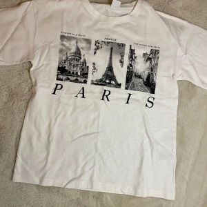 Vit t-shirt med Paris-motiv från Lindex - Säljer en vit t-shirt från Lindex med ett tryck av Paris-motiv. T-shirten har korta ärmar och är tillverkad i bomull. Perfekt för den som älskar Paris och vill ha en bekväm och stilren t-shirt. Använd en gång! Frakt ingår inte i priset, utan tas med köparen!