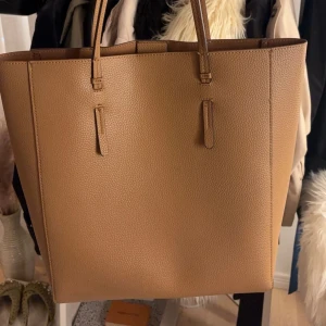 Beige shoppingväska - Snygg och rymlig beige väska. Perfekt för att bära med sig allt man behöver under dagen. Väskan har två långa handtag som gör den enkel att bära över axeln.