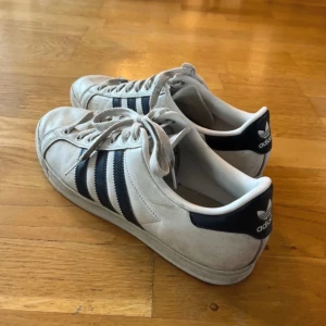 Adidas Superstar sneakers - Klassiska Adidas Superstar sneakers i vitt med svarta ränder. Skorna har snörning och den ikoniska tåhättan. Perfekta för en avslappnad stil.