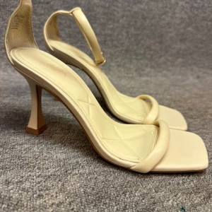 Beige klackskor med remmar - Eleganta beige klackskor med smala remmar och fyrkantig tå. Skorna har en stilren design med justerbart spänne runt ankeln och en hög klack för extra höjd.