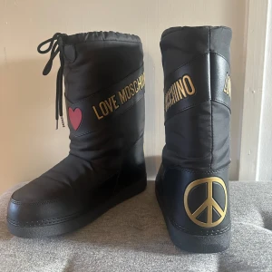 Svarta moon boots från Love Moschino - Snygga svarta moon boots från Love Moschino med guldtext och hjärtdetalj. Skorna har en rund tå och snörning upptill. På baksidan finns en stor fredssymbol i guld. Perfekta för vintern! Love Moschino Winter boots - nero  Aldrig använt, bara testat. Ångrat köp! Köpta för 1495kr.