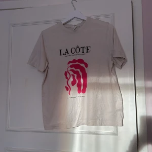 Beige t-shirt - Säljer en beige t-shirt med ett fint rosa tryck på framsidan. Texten 'Les Trouvailles des Plages' och 'Ode to Nature' ger en unik touch. Perfekt för en avslappnad stil.