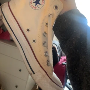 Vita sneakers från Converse - Klassiska vita Converse All Star sneakers med hög modell och snörning. Skorna har den ikoniska logotypen på sidan och en röd och blå rand längs sulan. Smutsiga, annars bra skick