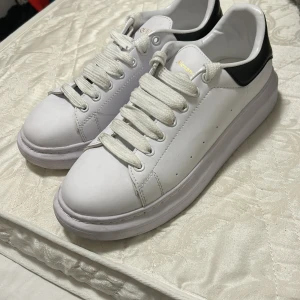 Vita sneakers från Alexander McQueen - Snygga vita sneakers från Alexander McQueen med svart detalj på hälen och guldfärgad logotyp. Skorna har klassisk snörning och är tillverkade i Italien.
