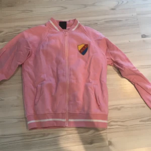 Rosa jacka med DIF-emblem - Säljer en rosa jacka med DIF-emblem på bröstet. Jackan har dragkedja framtill och långa ärmar. Den har ribbade muddar och krage med vita ränder. Perfekt för att visa ditt stöd för laget! Endast använd en gång annars helt ny från butiken. Nypris ungefär mellan 500-600 kr. 