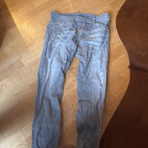 Ljusa jeans från Dsquared2 - Säljer ett par ljusa jeans från Dsquared2 med slitna detaljer och en patch på baksidan. De har en klassisk femficksdesign och en dragkedja. Perfekta för en avslappnad stil. De har ett ihop sytt hål mellan bena men det syns knappt när man använder dem 