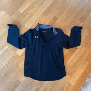 Svart jacka från Under Armour  - Säljer en svart jacka från Under Armour med dragkedja och långärmad design. Jackan har en lös passform och är perfekt för sportiga aktiviteter. Den har två praktiska fickor med dragkedjor framtill.